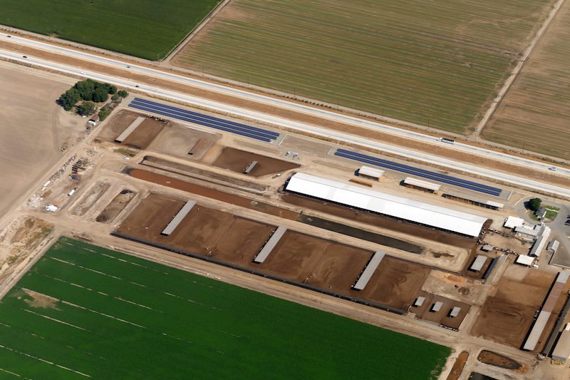 Aerial view of a small solar panel installation on a California dairy farm bordered by livestock pens, commercial farm buildings and four-lane highway.