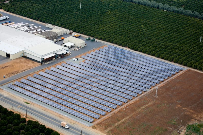 Aerial view of ground-mounted solar system situated next to commercial agricultural buildings and rows of orchard trees.
