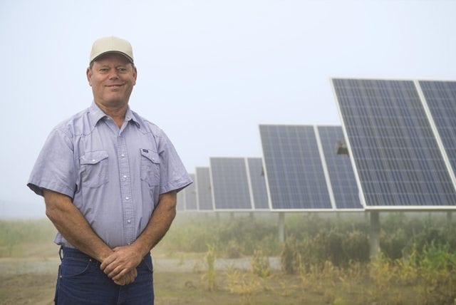 A farmer standing in front of rows of solar panels in a solar farm on California agricultural land.