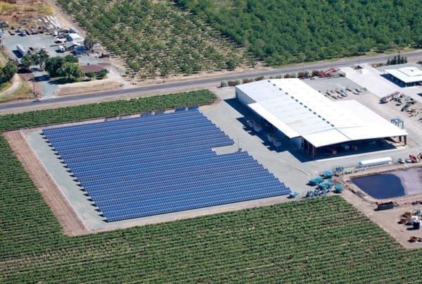 Aerial view of a solar installation on a farm in California surrounded by green crop fields, a commercial farm building, water retention pond and farm vehicles.