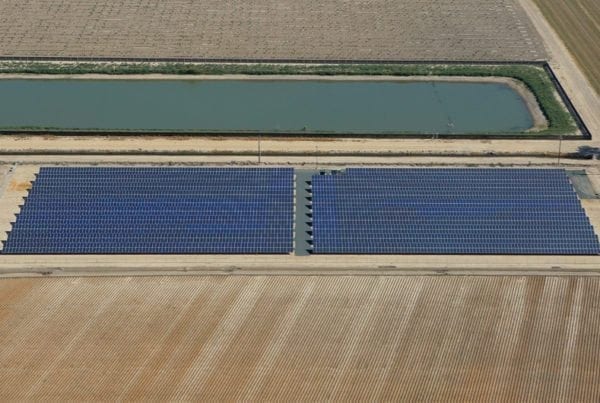 Aerial view of rows of solar panels in a solar installation on California agricultural land surrounded by plowed fields and a water retention pond.