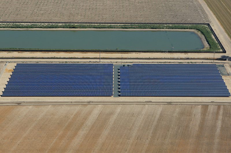 Aerial view of rows of solar panels in a solar installation on California agricultural land surrounded by plowed fields and a water retention pond.