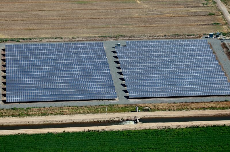 Horizontal rows of solar panels in a solar farm on agricultural land with green crops in the foreground.