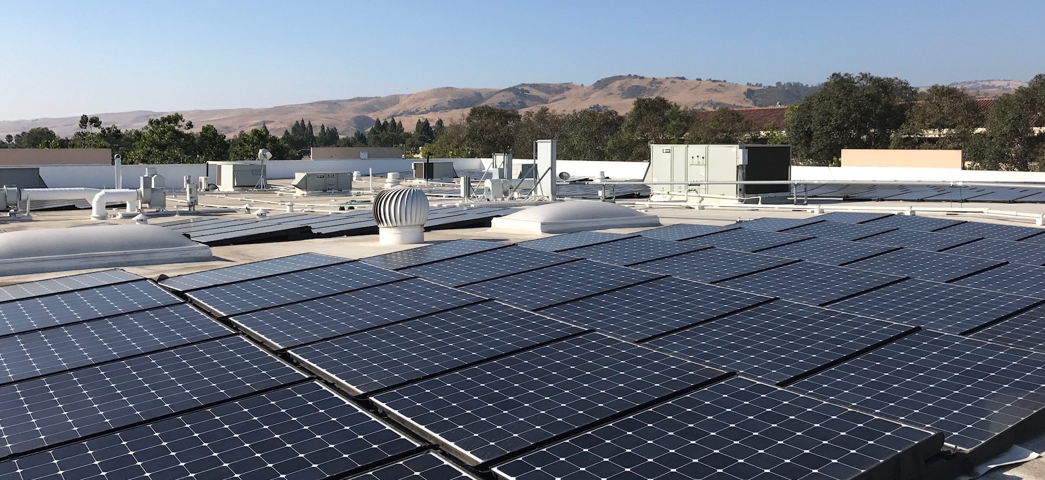 Looking across the roof of a commercial building with solar panels and trees and brown hills in the background.