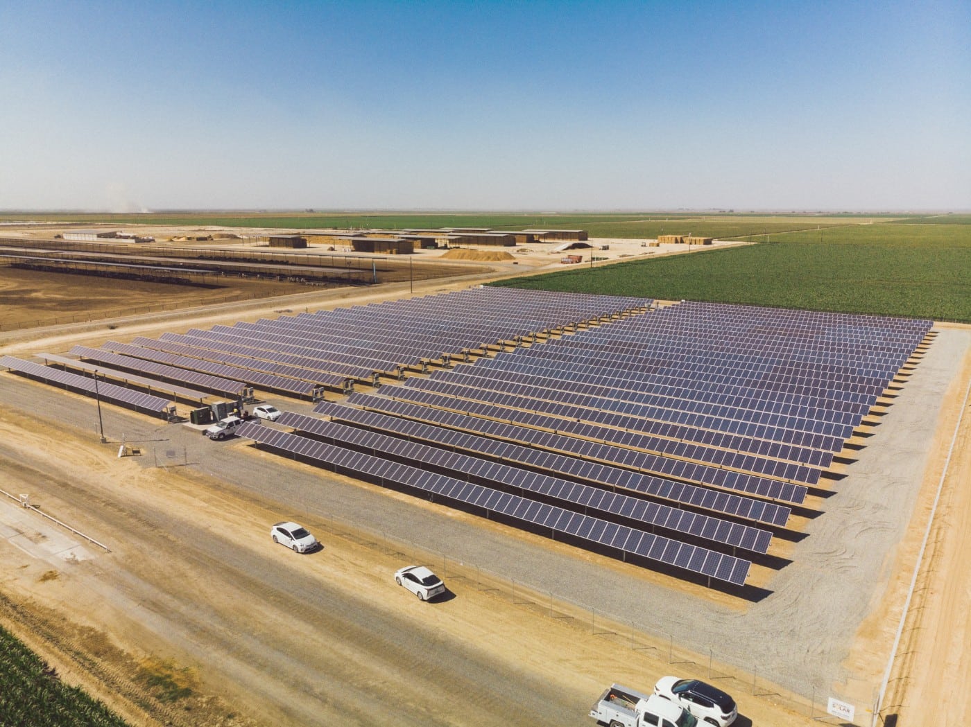 Aerial view of large ground-mounted solar panel installation at John Scheenstra Dairy surrounded by livestock pens, hay bales and green pastures in Wasco, California.