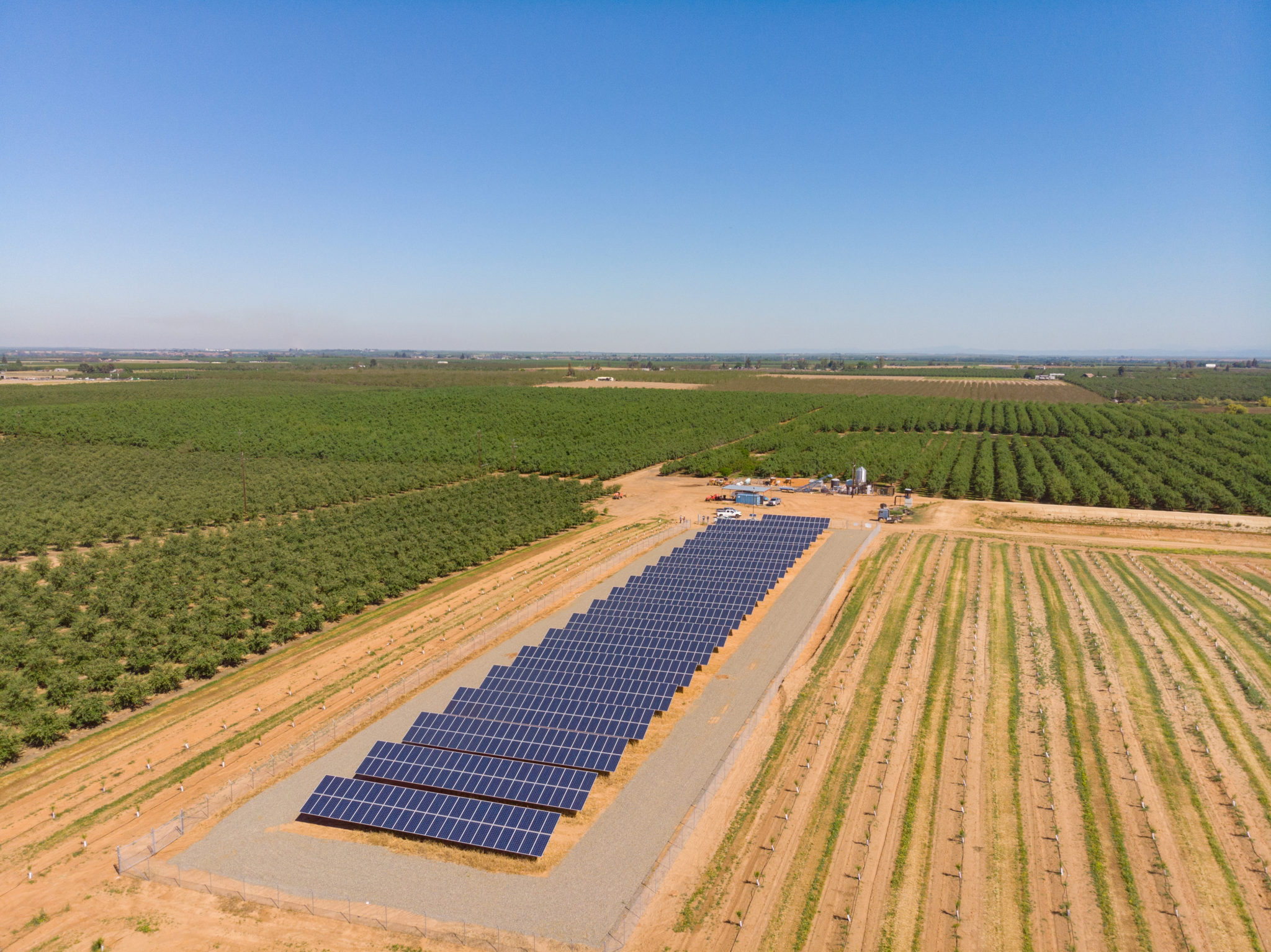 Aerial view of a small solar farm in Gustine, California with rows of crops in the background and a blue sky.