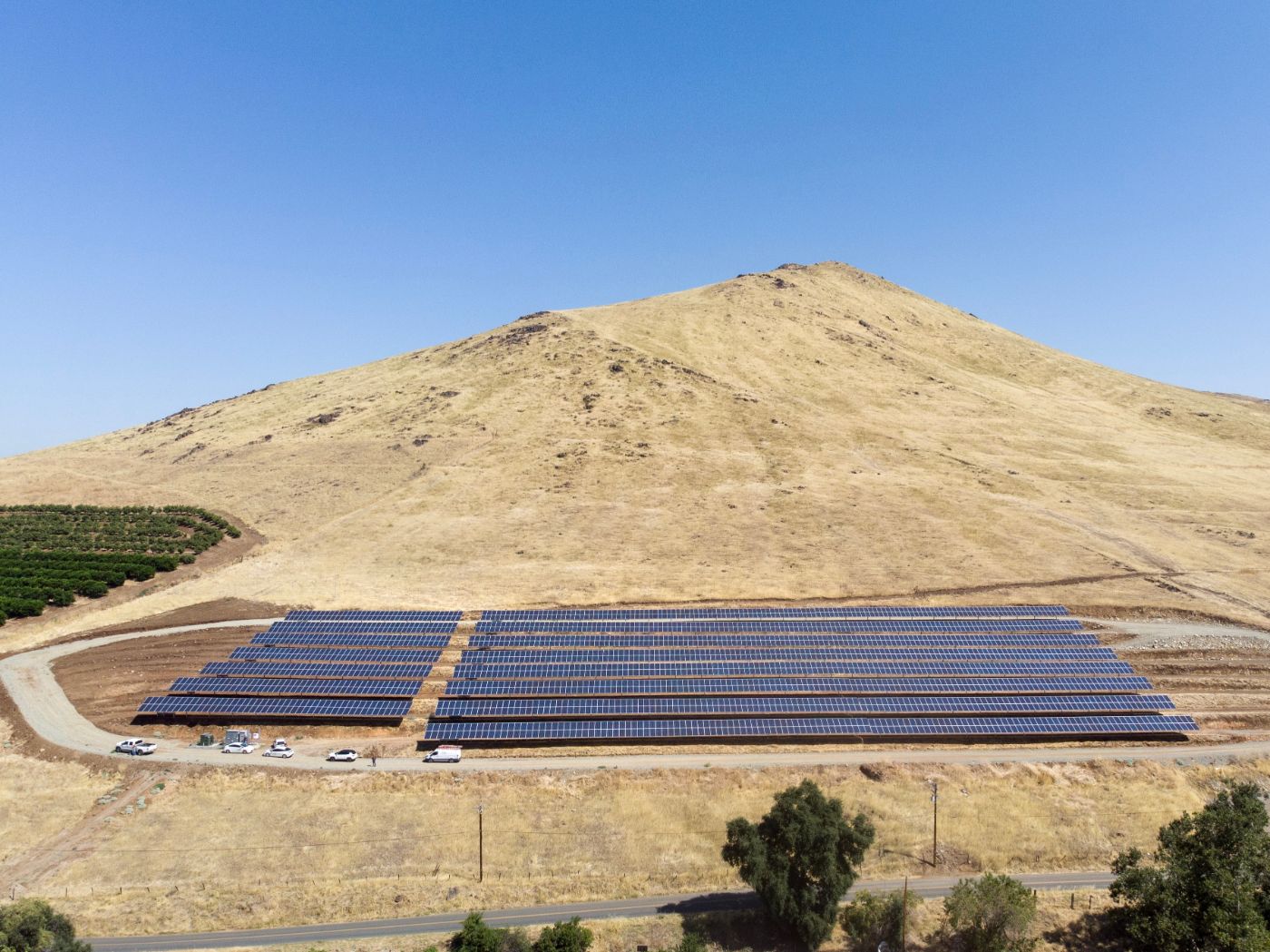 Aerial view of a small solar farm at the base of a brown hill with blue skies in the background.