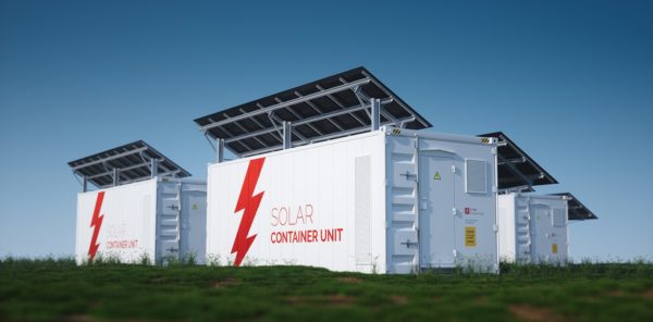 Several solar battery storage units sitting on green grass with roof-mounted solar panels against a blue sky.