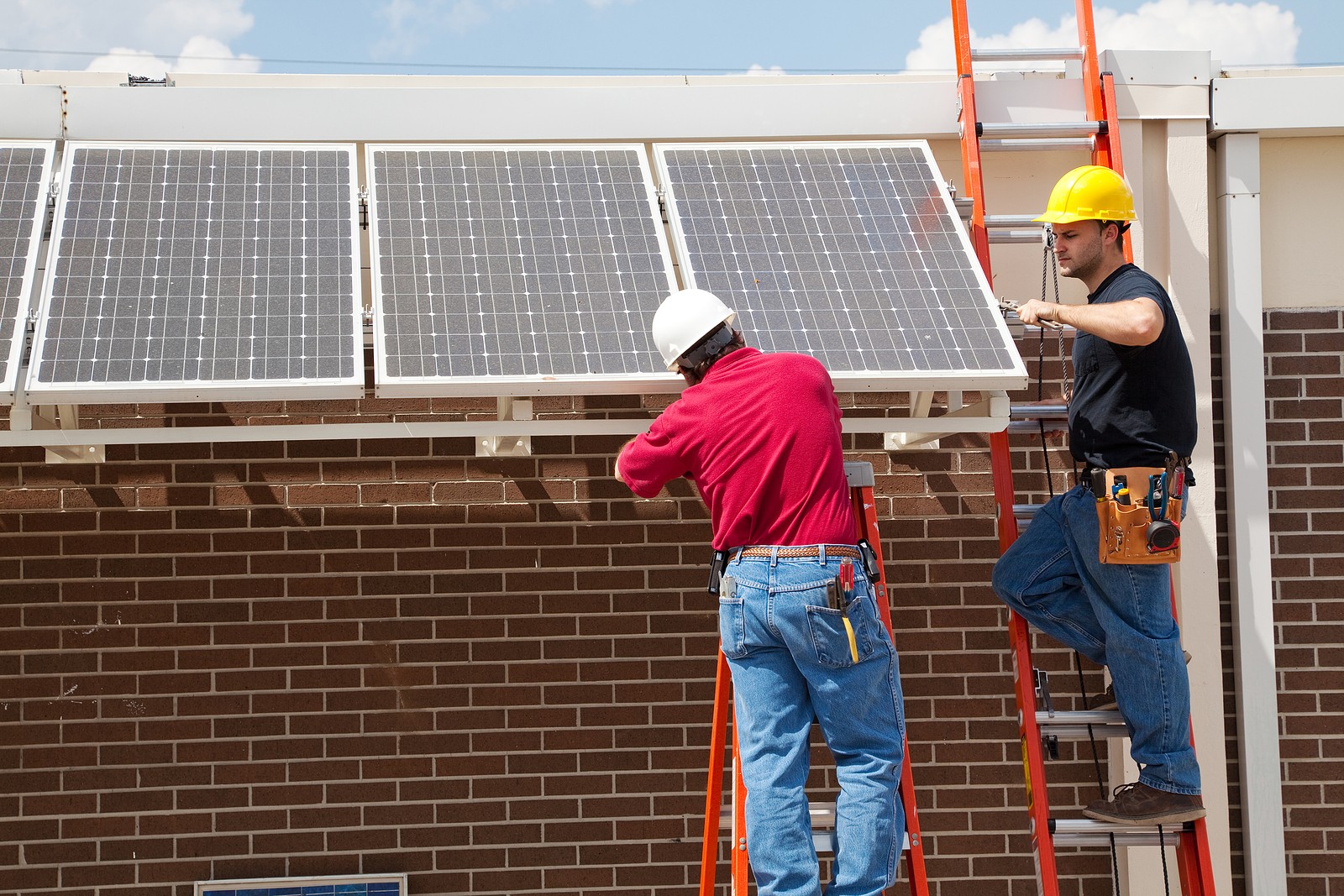 Two solar company employees installing solar panels on the side of a school building while standing on ladders.