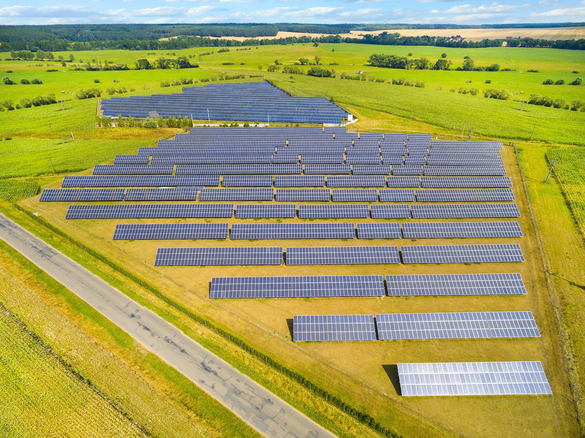 Aerial view of rows of solar panels in a small solar farm on flat green pastures with wooded areas in the background.
