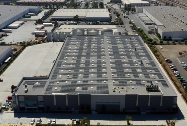 Aerial view of solar energy installation on the gray roof of a warehouse building in California with other warehouses in the background.