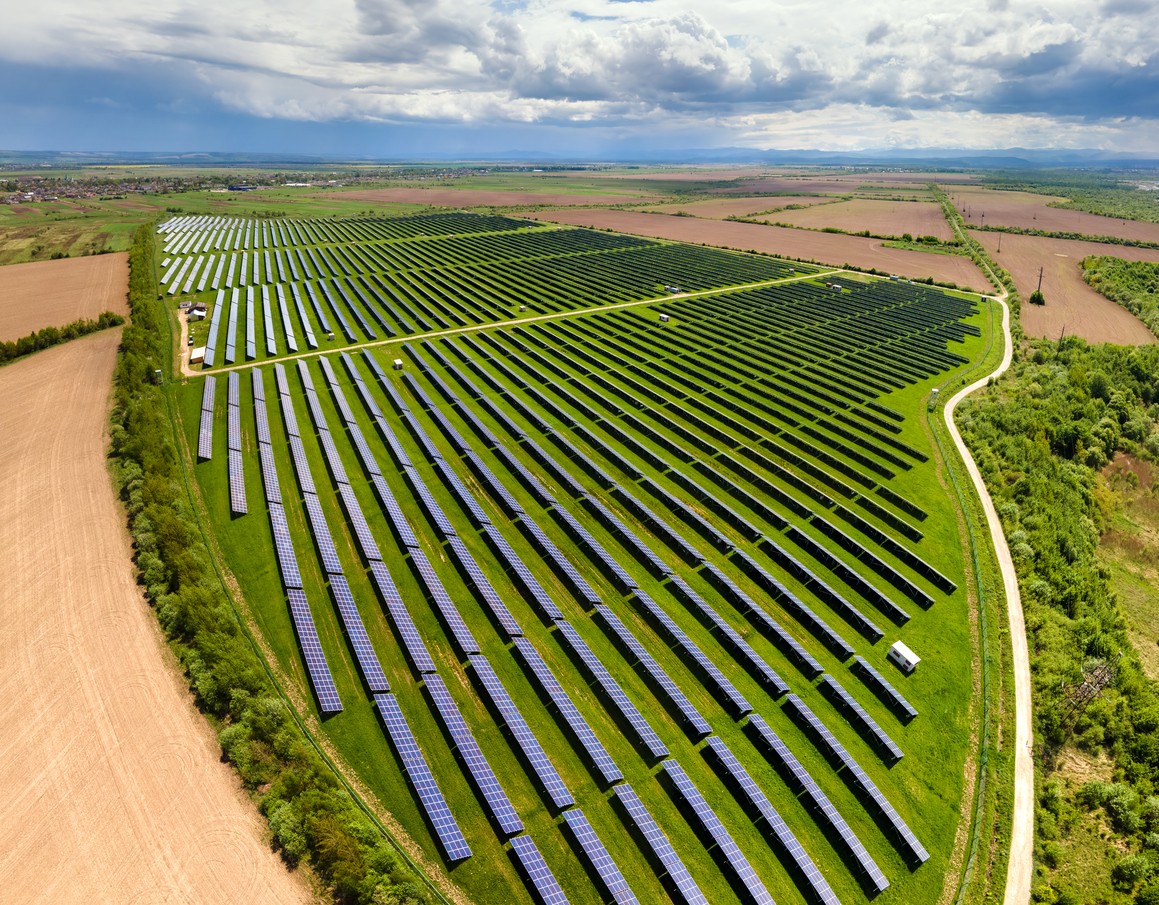 Aerial view of a large solar farm on green land surrounded by plowed brown fields and a cloudy sky.