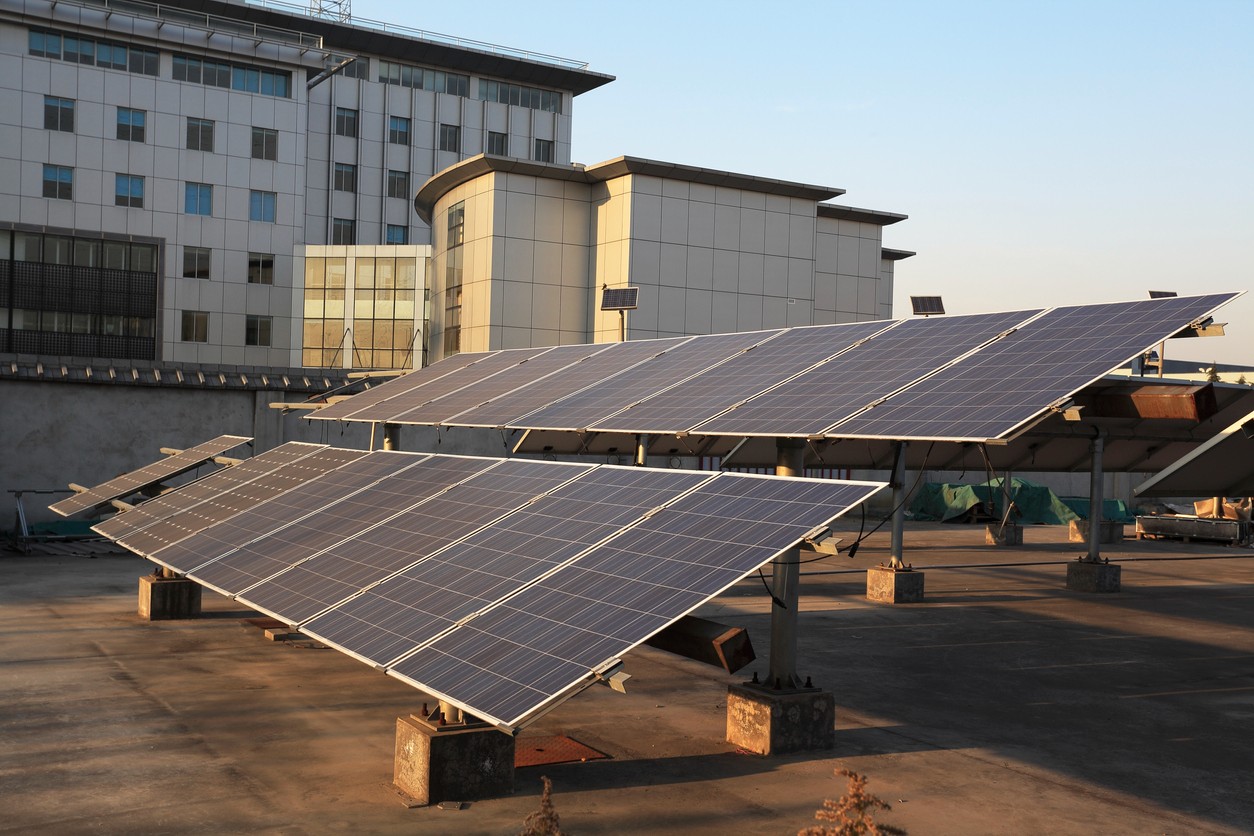 Two rows of solar panels mounted on the roof of a commercial building with an office building in the background.