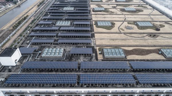 Aerial view of rows of solar panels on the left side of the roof of an industrial plant.