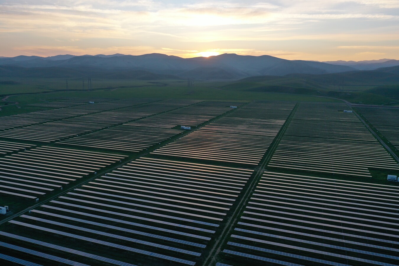 A large utility-scale solar farm with rows of solar panels against green mountains at dusk.