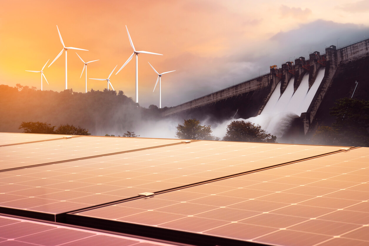 Solar panels in the foreground with wind turbines and a hydro plant in the background at sunset.