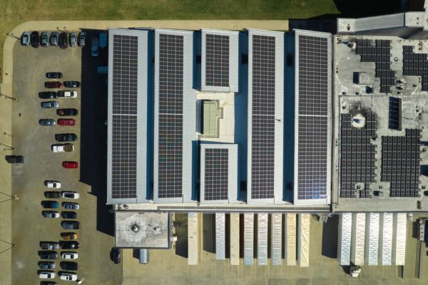Aerial view of black solar panels on the roof of a warehouse with trucks parked at loading docks and cars in a parking lot.