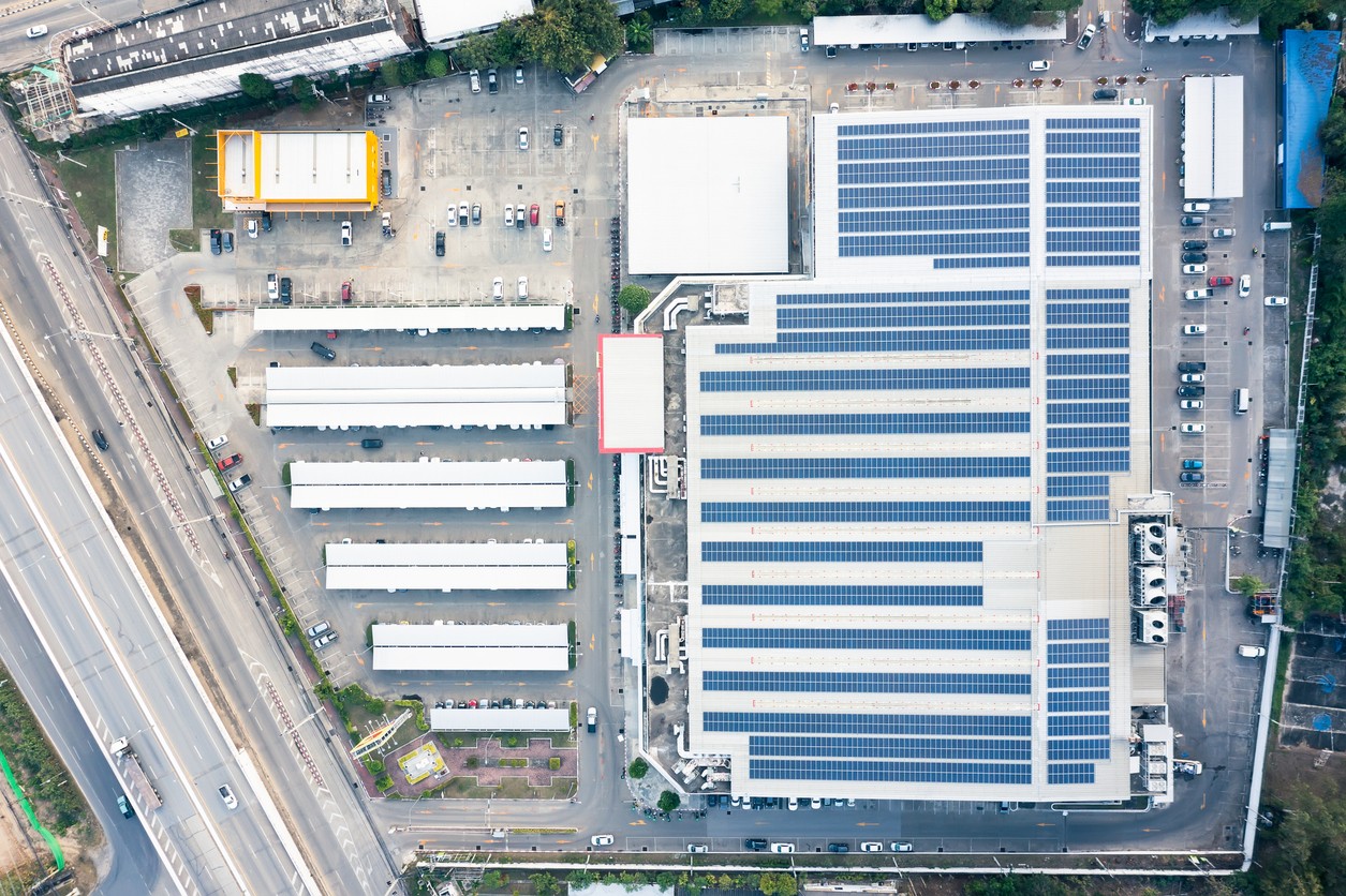 Aerial view of a warehouse with blue solar panels and covered parking spaces alongside a highway.