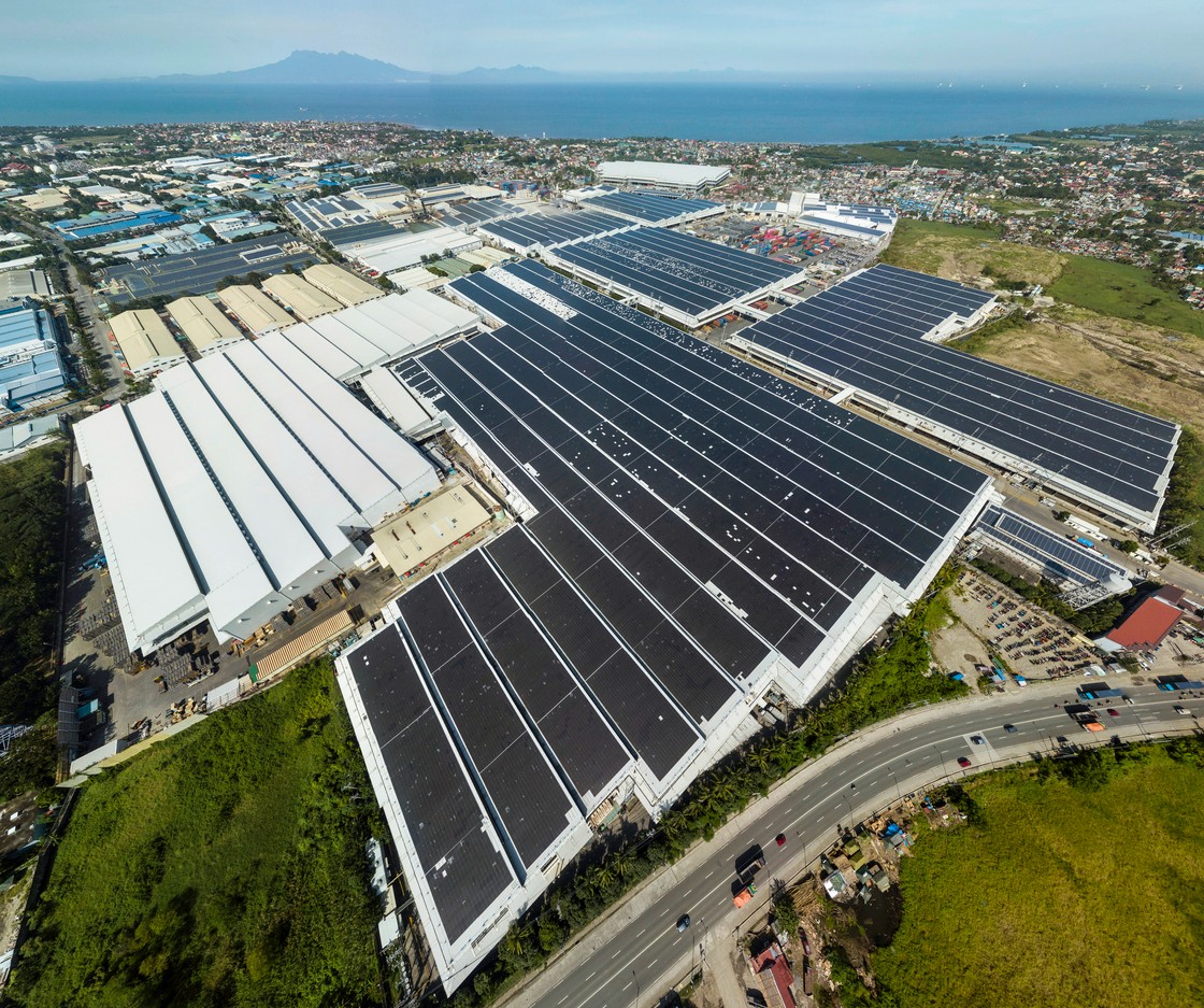 Aerial view of rows of black solar panels on the roofs of a cluster of warehouses with ocean, mountains and sky in the background.