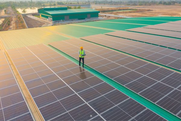 Aerial view of a solar company worker standing among black commercial solar panels on a green building roof with a green building in the background.