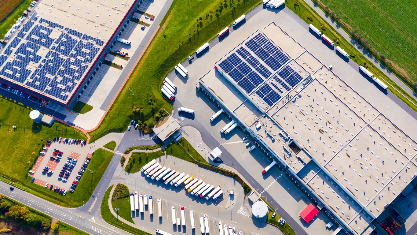 Aerial view of two warehouses with blue solar panels on the roofs and trucks parked at loading docks around the buildings.