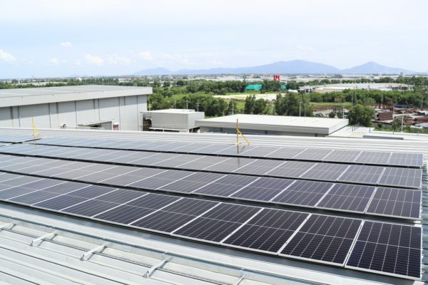 Four rows of solar panels on a commercial building roof with green landscaping and mountains in the background.