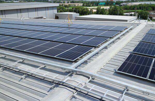 Rows of solar panels mounted on the roof of an industrial building with green landscaping and trees in the background.