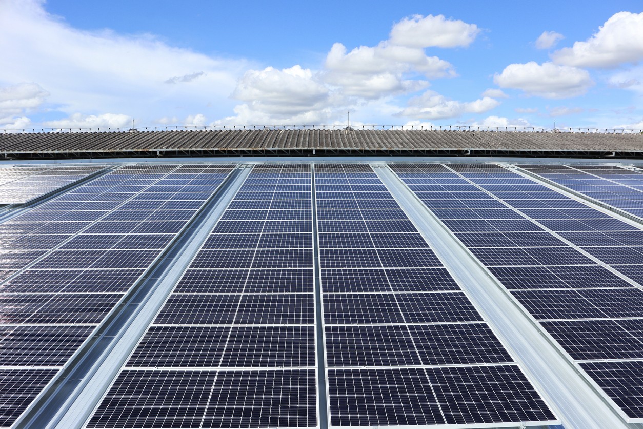 Looking across rows of black solar panels on an industrial building rooftop with a blue sky and clouds.