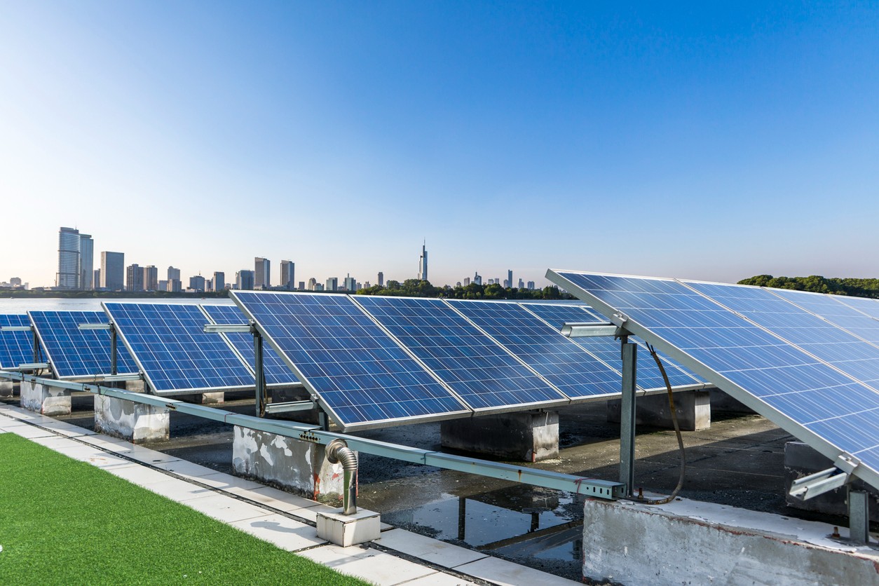 Rows of ground-mounted blue commercial solar panels with a blue sky and city skyline in the background.
