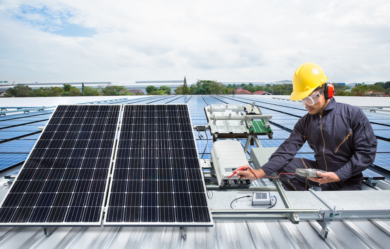 Solar company engineer in a yellow hardhat testing a commercial solar panel on the roof of a commercial building.