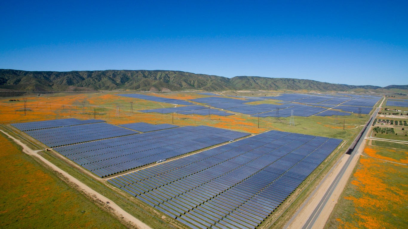 Aerial view of a large utility-scale California solar farm surrounded by poppy fields and green mountains in the background.