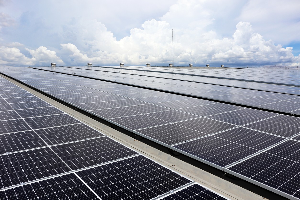 Closeup view of rows of black solar panels on the roof of an industrial building with cloudy skies in the background.