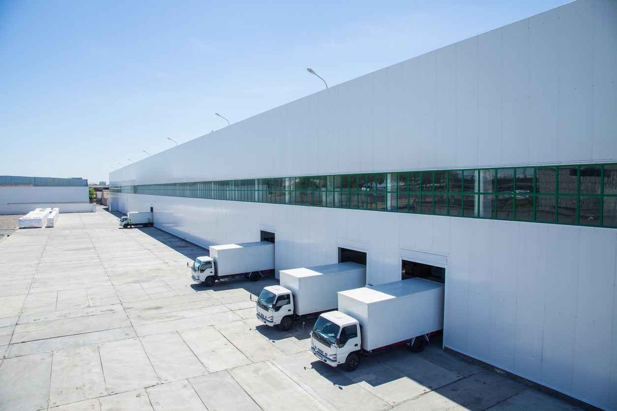 Four white semi trucks parked at loading docks along the side of a modern white warehouse building with glass windows.
