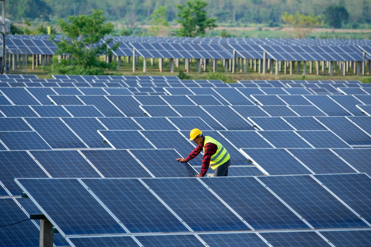 A solar company engineer in the middle of rows of solar panels with another solar installation and greenery in the background.