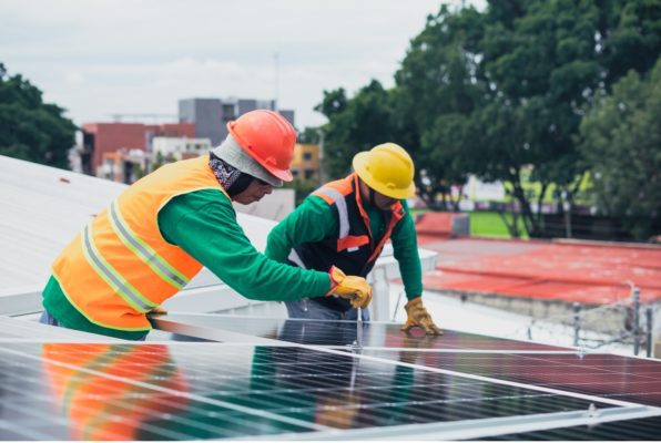 Two solar company workers in hardhats installing a solar panel on a commercial building roof.