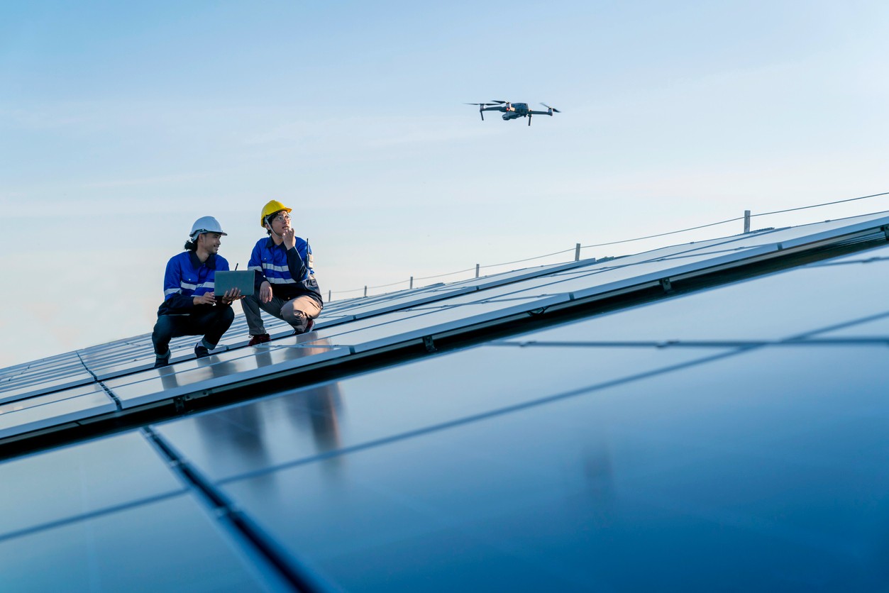 Two solar company workers squatting on a roof operating and watching a drone fly over commercial solar panels.