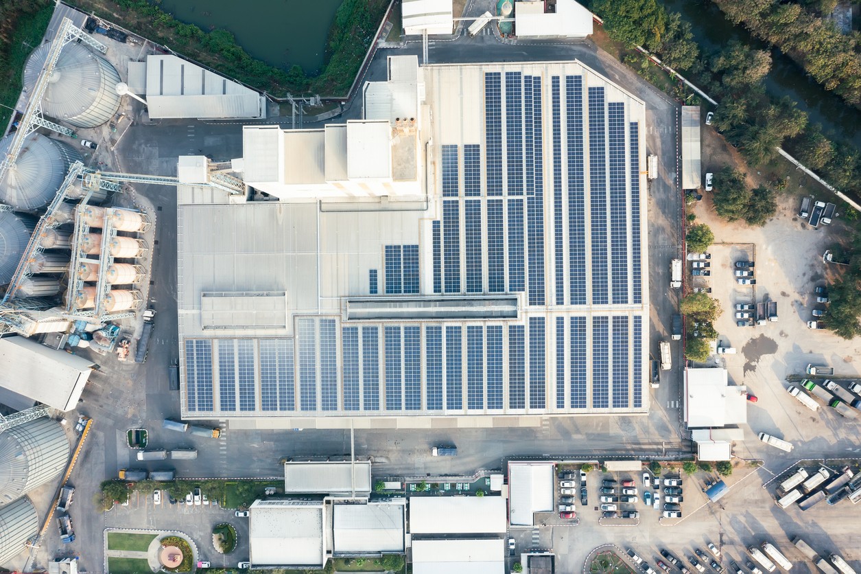 Aerial view of rows of solar panels on the roof of an industrial plant with smaller buildings and cars parked around it.