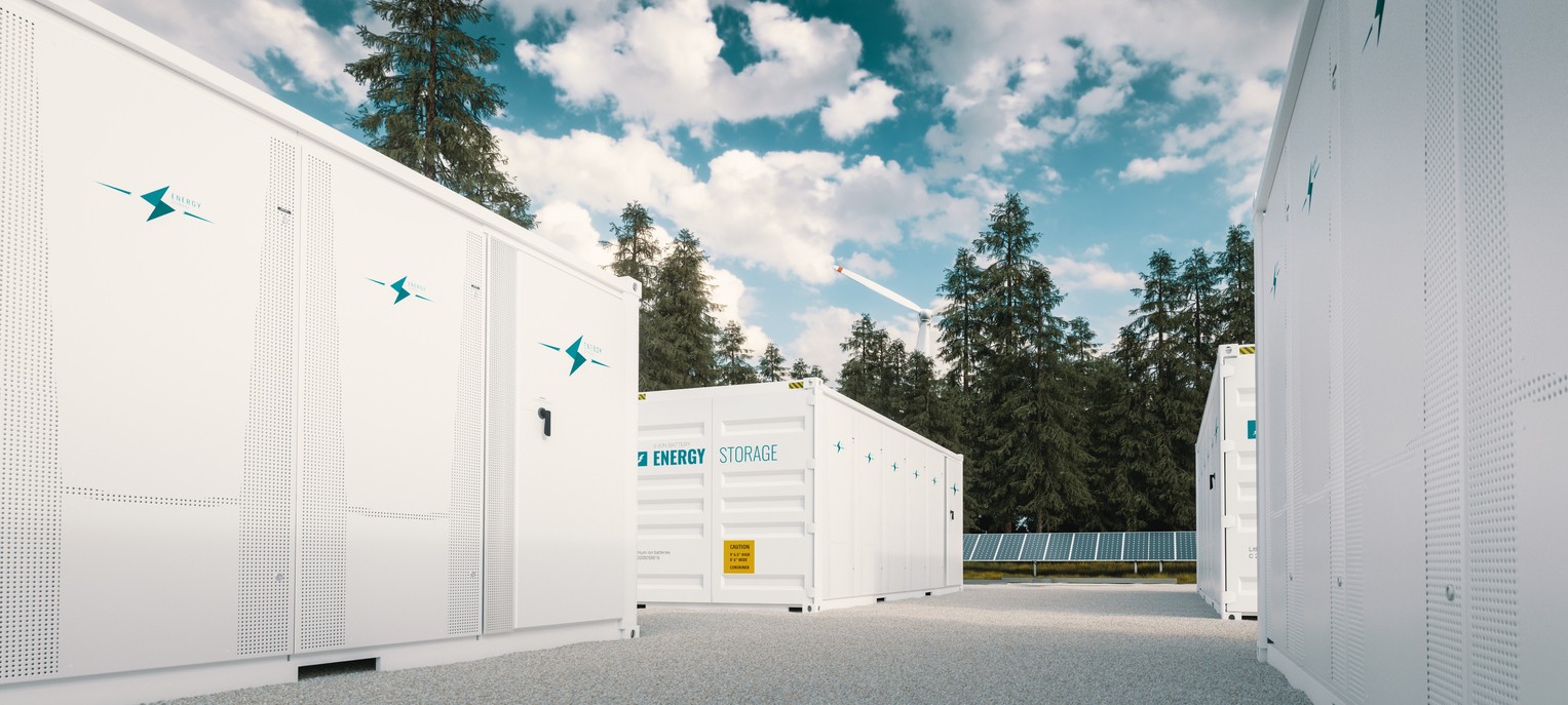 Close up view of solar battery storage units with ground-mounted solar panels and a wind turbine in the background.