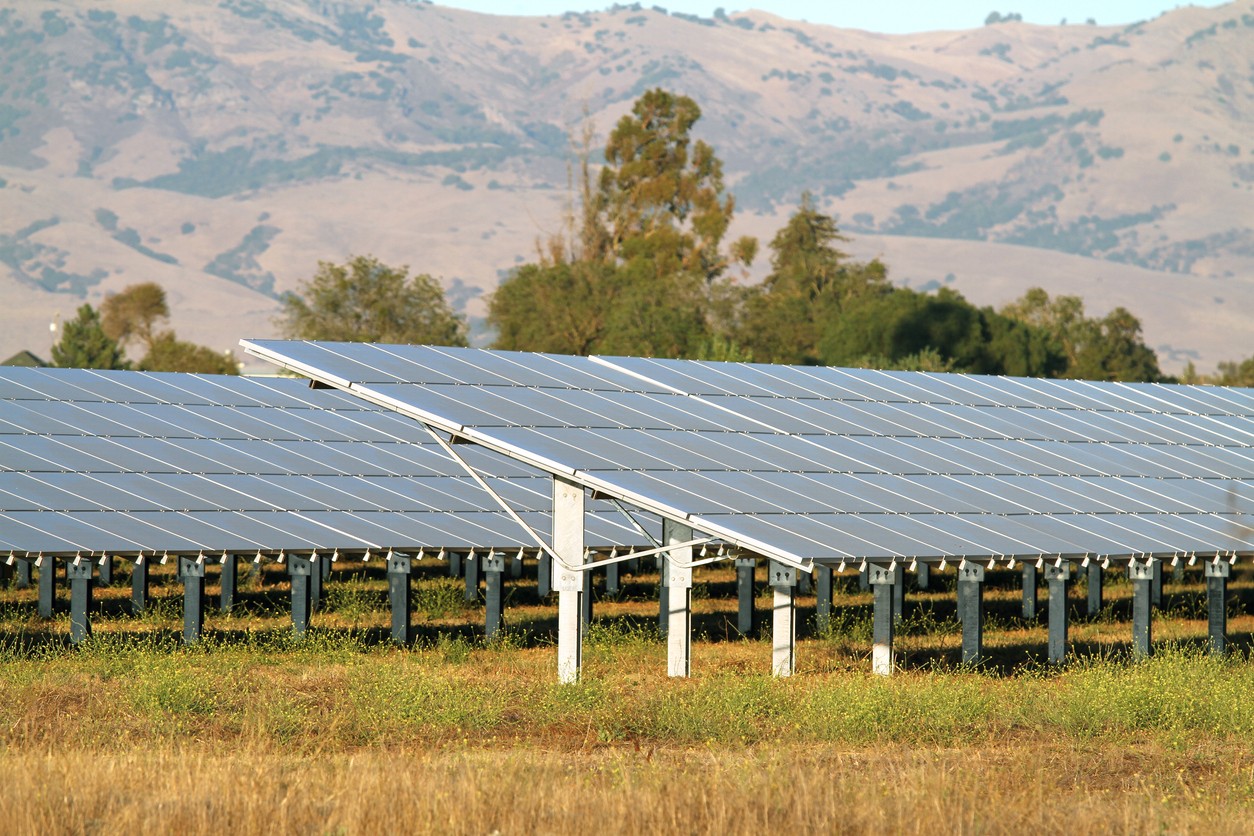 Rows of large ground-mounted solar panels against trees and California mountains in the background.
