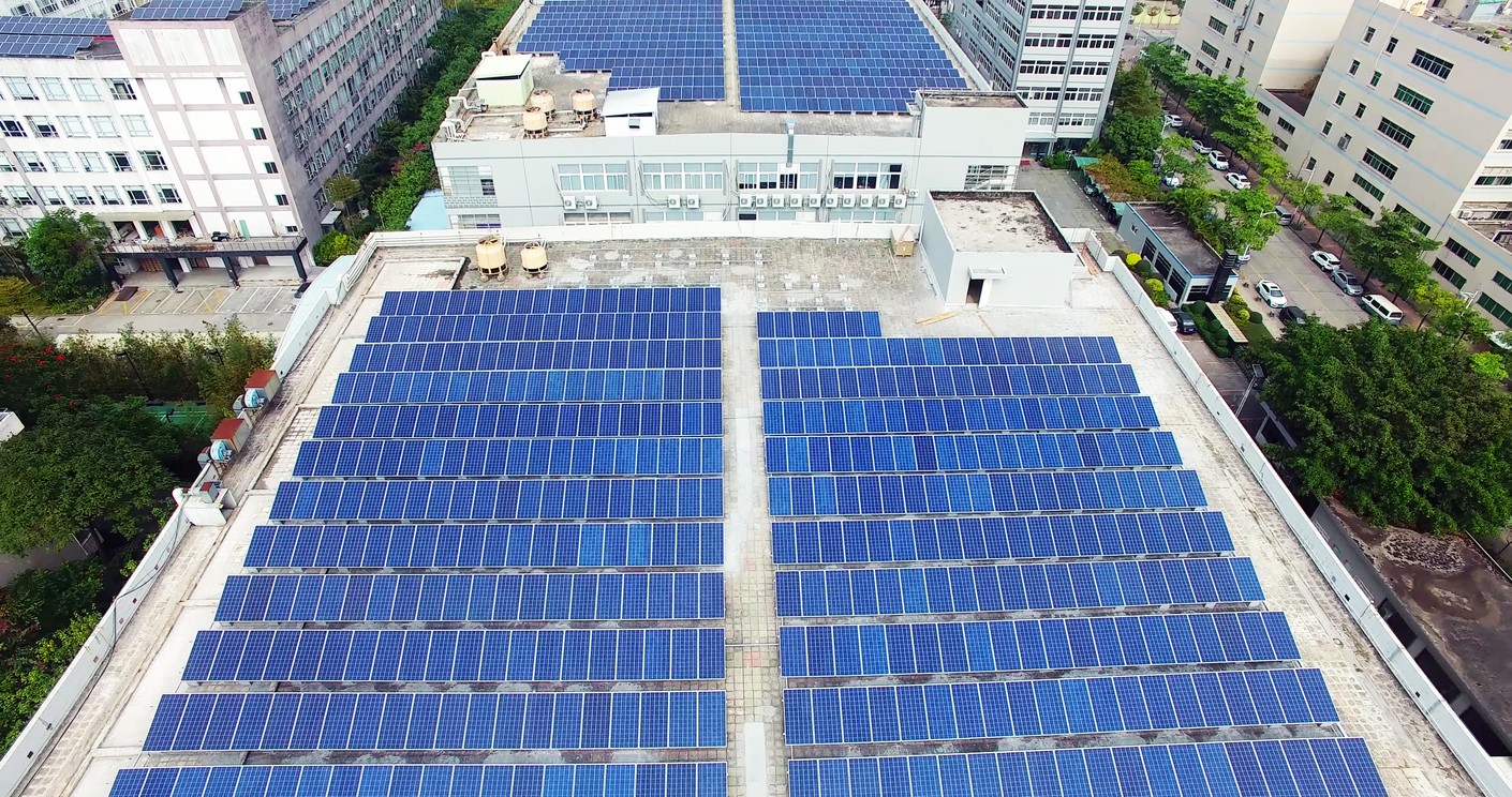 Looking down on rows of solar panels on the roof of an office building among other office buildings in a city.