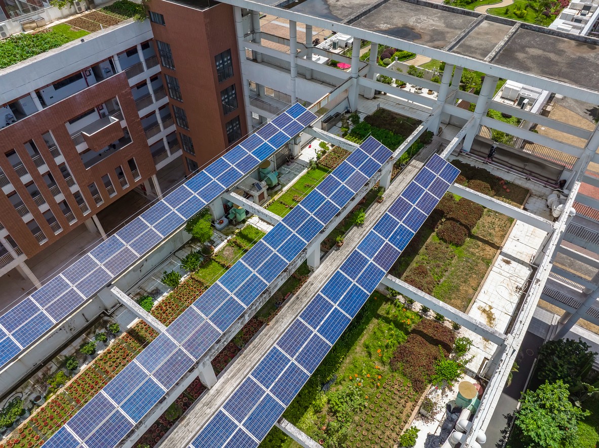 Aerial view of three rows of solar panels and a community garden on the roof of a school building.