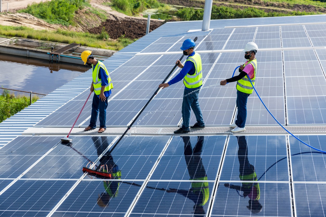 Three solar company workers cleaning solar panels with a water hose and mops on the roof of a commercial building.