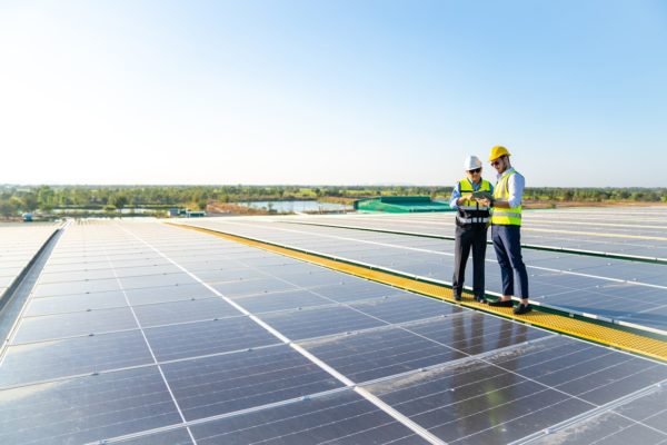 Two solar company workers standing on a commercial building roof between rows of solar panels and examining a computer tablet.