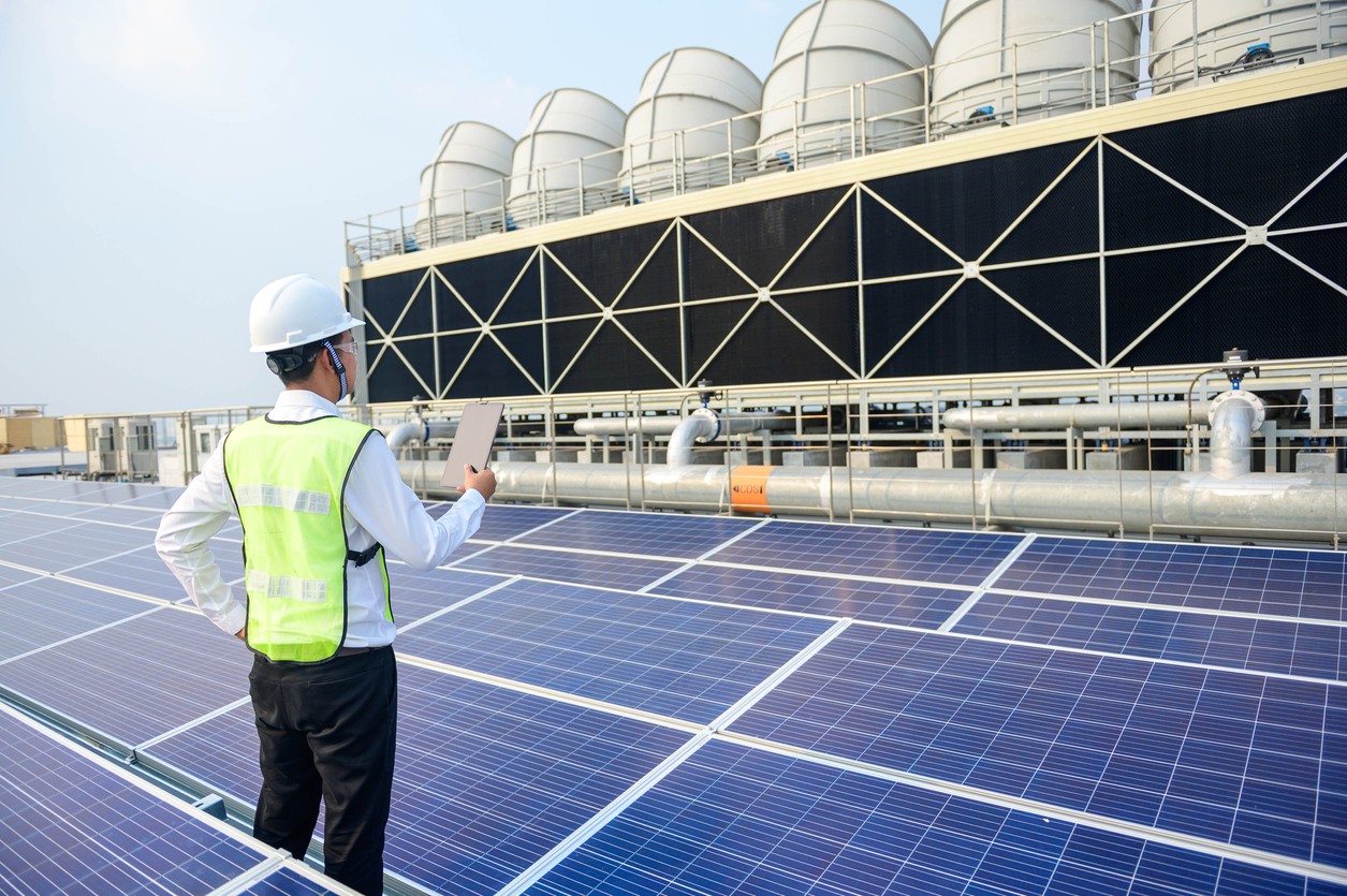 Solar company worker on the roof of an industrial building inspecting solar panels with large pipelines and structures in the background.