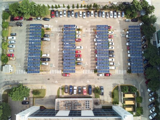 Aerial views of four rows of solar carports in a parking lot with cars parked underneath them. 