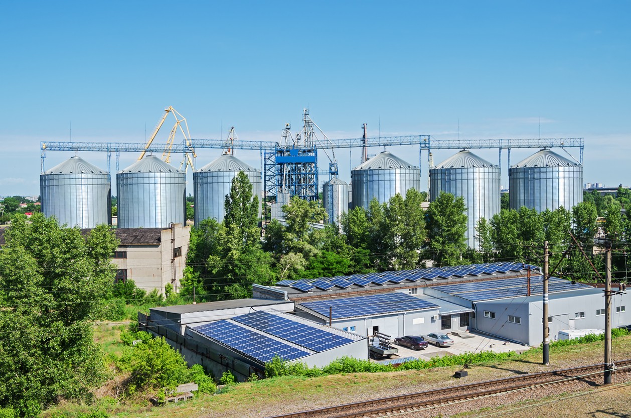 Solar panels on roof of a commercial grain mill building with trees, grain silos and blue sky in the background.