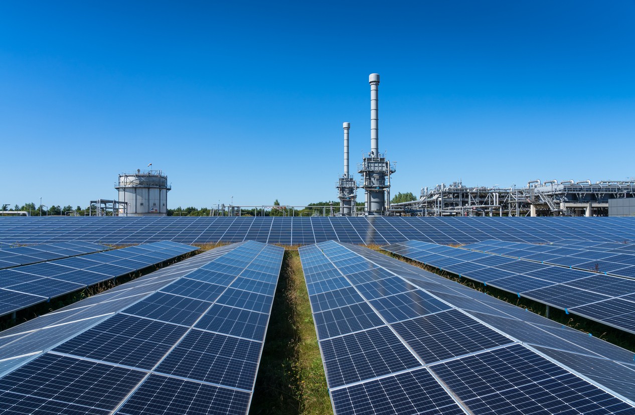 Rows of solar panels on the roof of an industrial plant with industrial smokestacks in the background.