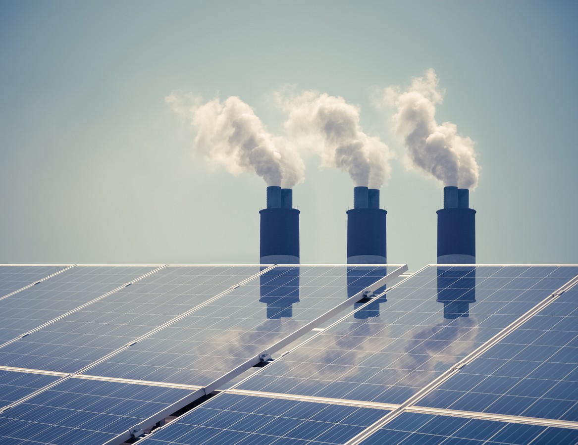 Closeup view of commercial solar panels with three industrial smokestacks in the background.